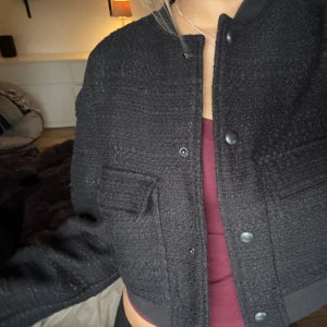 Svart bomberjacka från Nelly XS - Svart bomberjacka från Nelly i storlek XS. Jackan är croppad med långa ärmar, har två stora fickor framtill och stängs med knappar. Materialet är mjukt och strukturerat, med ribbade muddar vid ärmslut och nederkant. Perfekt för dig som gillar en chill och trendig look.