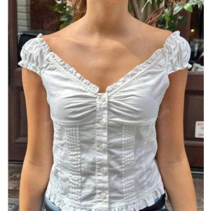 Vit blommig blus med volang och knappar - Supersöt vit blus med broderade blommor, volangkant vid hals och ärmar samt knappar framtill. Blusen har v-ringning, korta puffärmar och är lätt croppad. Perfekt för sommarens alla tillfällen, och materialet är mjuk bomull med spetsdetaljer.