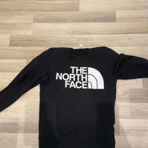 Svart sweatshirt från The North Face - Svart sweatshirt från The North Face med stor vit logga framtill. Klassisk rund halsringning och långa ärmar. Tillverkad i mjuk bomullsmix som är skön att ha på sig. Perfekt för dig som gillar streetwear och enkel stil.