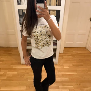Vit leopardmönstrad t-shirt från Zara - Vit t-shirt från Zara med coolt leopardmönster i guld och svart framtill. Trycket har texten 'SPIAGGIA DELLA PELOSA' ovanför. Klassisk passform med korta ärmar och rund hals. Perfekt för dig som vill sticka ut lite extra.