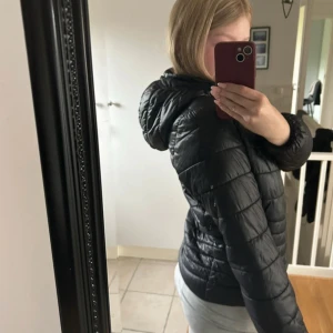 Svart pufferjacka med huva - Säljer en svart pufferjacka med huva och dragkedja framtill. Jackan har quiltad design och två fickor med dragkedja. Perfekt för kyligare dagar och enkel att matcha med olika outfits.