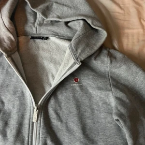 Grå hoodie från Björn Borg - Mysig grå hoodie från Björn Borg med dragkedja framtill, huva och två fickor. Är en xs men passar S Liten röd logga på bröstet och mjukt fleecefoder på insidan. Perfekt för chill dagar eller när du vill vara extra bekväm.