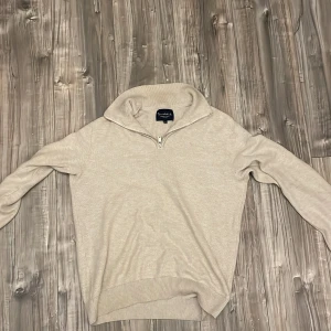 Beige stickad half zip-tröja Bondelid - Snygg beige stickad tröja från Bondelid med half zip-dragkedja och hög krage. Tröjan har lång ärm och diskret broderad logga på bröstet. Perfekt för lager-på-lager och chill stil.
