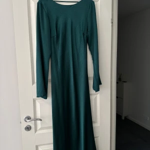 Mörkgrön långklänning med knyt - Snygg långklänning i mörkgrönt med lång ärm och rund halsringning framtill. Baksidan har en djup v-ringning och ett band. Enbart använd då gånger.