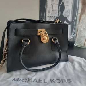 Svart handväska från Michael Kors, Hamilton  - Stilren svart handväska från Michael Kors i skinn med guldfärgade detaljer, kedjerem och klassiskt MK-lås framtill. Väskan har både handtag och axelrem, samt dragkedja på insidan. Perfekt för dig som vill ha en snygg och praktisk väska med lyxig känsla.