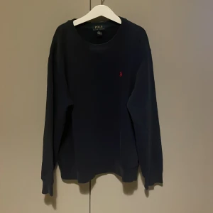 Mörkblå tröja från Polo Ralph Lauren - En stilren mörkblå tröja från Polo Ralph Lauren med klassisk rund hals och röd broderad logga på bröstet. Tröjan har långa ärmar och är tillverkad i mjuk bomull, perfekt för lager-på-lager under kyliga dagar | priset kan alltid diskuteras!