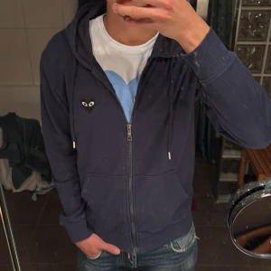 Mörkblå hoodie från Comme des Garçons - Säljer en mörkblå hoodie från Comme des Garçons Play med dragkedja och huva. Ikoniskt hjärta med ögon på bröstet och stort hjärta med ögon på ryggen. Skön passform och snörning i huvan. Perfekt för dig som gillar streetwear vibes.