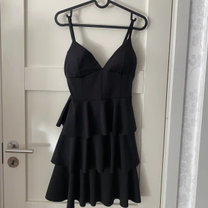 Svart volangklänning med smala axelband - Supersnygg svart kort klänning med flera volanger och smala justerbara axelband. Klänningen har v-ringad urringning och figurnära passform upptill. Perfekt för dig som vill sticka ut med en trendig och lekfull look.