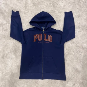 Marinblå hoodie från Polo Ralph Lauren - Säljer en marinblå hoodie från Polo Ralph Lauren med dragkedja, stor huva och känguruficka. Framsidan har stor orange POLO-logga och ärmen har broderad röd polospelare. Mjuk bomullsblandning, perfekt för chill dagar.