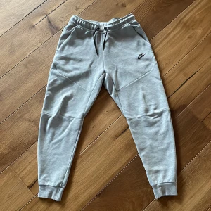 Grå mjukisbyxor från Nike med snörning - Grå mjukisbyxor från Nike med svart logga på vänster ben, elastisk midja med svart snörning och ribbade muddar vid bensluten. Byxorna har fickor på sidorna och är tillverkade i mjukt bomullsmaterial för maximal komfort.