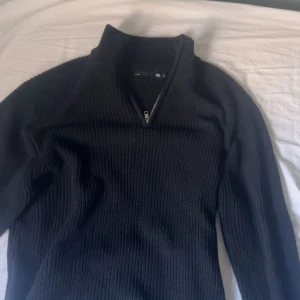 Svart ribbad stickad tröja half zip - Svart ribbstickad tröja med hög krage och half zip-dragkedja framtill. Långärmad och i ett mjukt, stickat material som passar perfekt när det är kyligt. Enkel och stilren design som är lätt att matcha.