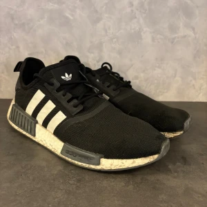 Adidas NMD  - Svarta Adidas NMD sneakers med vita detaljer och klassiska tre ränder på sidan. Ovandel i mesh och syntet, vit boost-sula och svarta snören. Skorna har en sportig vibe och är perfekta för dig som gillar streetwear. Adidas-logga på plös och häl.