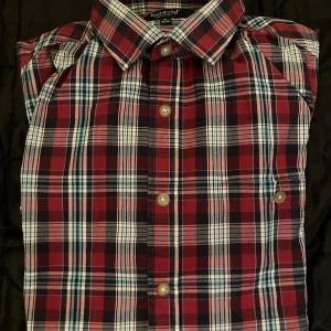 Rutig röd skjorta från Redwood - Snygg rutig skjorta från Redwood i rött, blått, vitt och svart. Klassisk krage, långa ärmar och knappar framtill. Perfekt för dig som gillar en chill och stilren look. Skjortan är gjord i mjuk bomull och har en normal passform.