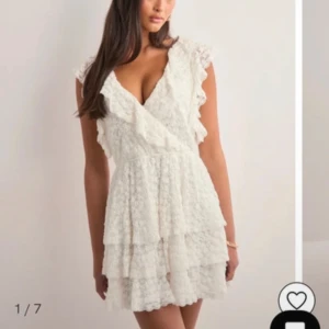 Vit spetsklänning med volanger - Supersöt vit kortklänning med omlott och v-ringad hals, volangdetaljer på axlar och kjol, samt öppen rygg med knytband. Klänningen har flera lager och är gjord i spets med blommigt mönster. Perfekt för sommarens alla tillfällen! Klänning har använts endast 1 gång! Ny pris: 600kr.