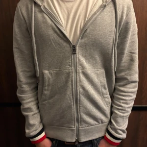 Grå Moncler hoodie med dragkedja - Snygg grå hoodie från Moncler med dragkedja, huva och klassiska Moncler-loggan på ärmen. Tröjan har ribbade muddar med röd, vit och marinblå rand samt matchande detalj på huvan. Perfekt för dig som gillar stilrena och sportiga plagg.