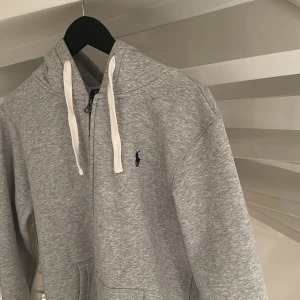 Grå hoodie från Polo Ralph Lauren M - Snygg gråmelerad hoodie från Polo Ralph Lauren med dragkedja, vit snörning i huvan och klassisk broderad logga på bröstet. Mjuk insida och känguruficka framtill. Perfekt för chill dagar eller när du vill ha en clean look.