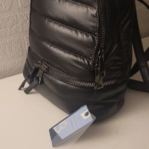  Helt ny Svart quiltad ryggsäck i syntet -  Helt ny Stilren svart ryggsäck i  design med blanka ytor. Väskan har dubbla dragkedjor med snygga detaljer och justerbara axelremmar. Tillverkad i syntetmaterial som ger en modern look och passar perfekt till din vardagsstil. 30×30×10