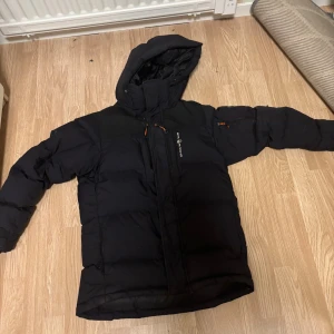 Svart dunjacka med huva från Sail racing  - Säljer en svart dunjacka från Sail Racing med huva och flera dragkedjeförsedda fickor. Jackan är tjockt vadderad och har en stilren look med diskreta loggor. Perfekt för kalla dagar och har lång ärm samt hög krage för extra värme.