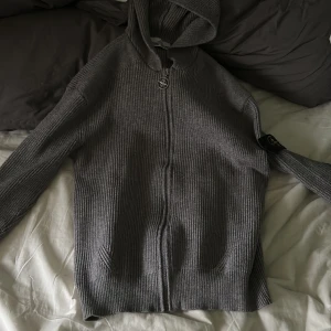 Grå stickad hoodie från Stone Island - Riktigt snygg grå stickad hoodie från Stone Island med dragkedja och ikonisk patch på ärmen. Tröjan har huva, ribbad struktur och långa ärmar. Perfekt för dig som vill ha en stilren och exklusiv look med streetkänsla.