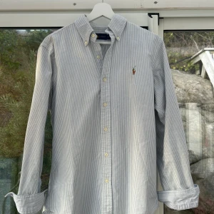 Randig skjorta från Ralph Lauren - Snygg skjorta från Ralph Lauren med smala blå och vita ränder, klassisk button-down krage och broderad logga på bröstet. Långärmad modell i mjuk bomull med normal passform och knappar framtill. Perfekt för dig som gillar stilrena och tidlösa plagg.