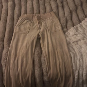 Beige mjukisbyxor Juicy Couture S - Supermjuka beige mjukisbyxor från Juicy Couture i velour med bred resår och snörning i midjan. Stora fickor fram och bak med knappdetaljer. Perfekta för en chill och trendig look.