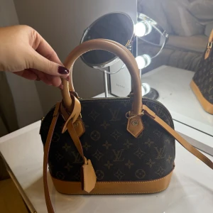 Handväska brun - Snygg handväska från  i klassiskt monogrammönster. Väskan är brun med ljusbruna skinnhandtag och detaljer, har dragkedja upptill och guldiga metalldetaljer. Perfekt storlek för det viktigaste och en riktig ikon i garderoben.