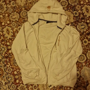 Vit hoodie från Sail Racing med dragkedja - Vit hoodie från Sail Racing med broderad logga på bröstet och huva. Tröjan har hel dragkedja framtill men därmot har en sönder kedja vid bröstet men går o dra med händerna !, ribbade muddar och är tillverkad i en mjuk bomulls- och polyestermix. Perfekt för chill dagar eller när du vill ha en clean look.