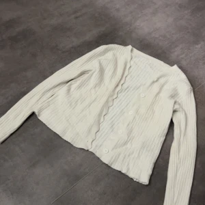 Vit ribbad kofta med vågig kant - Säljer en söt vit kofta med ribbat tyg och lång ärm. Koftan har knappar framtill och en vågig kant längs öppningen som ger en extra detalj. Inte använd så mycket, som ny nästan!