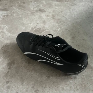Svarta fotbollsskor från Puma - Säljer ett par svarta fotbollsskor från Puma med vita detaljer och klassisk snörning. Skorna har rund tå och platt sula med fasta dobbar för bra grepp på planen. Tillverkade i syntetmaterial och designade för att ge en sportig look.