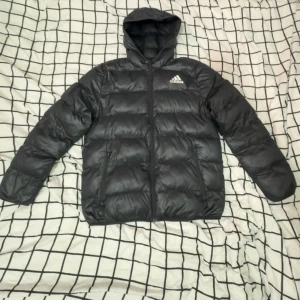 Svart pufferjacka från Adidas - Svart pufferjacka från Adidas med huva och dragkedja framtill. Jackan har två sidofickor med dragkedja och klassisk Adidas-logga på bröstet. Perfekt för kalla dagar med sin vadderade design och sportiga look.