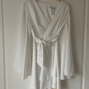 Vit omlottklänning från Adoore - Superfin vit kort omlottklänning från Adoore med långa vida ärmar och knyt i midjan. Klänningen har v-ringning och ett flowy fall som ger en romantisk vibe. Perfekt för sommarens alla tillfällen!