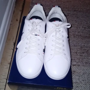 Vita sneakers Polo Ralph Lauren strl 47 - Snygga vita sneakers från Polo Ralph Lauren med marinblå detaljer och klassisk logga på sidan. Skorna har rund tå, snörning och platt sula. Materialet är en mix av skinn, syntet och textil med gummisula. Perfekt för dig som gillar stilrena och sportiga sneakers.