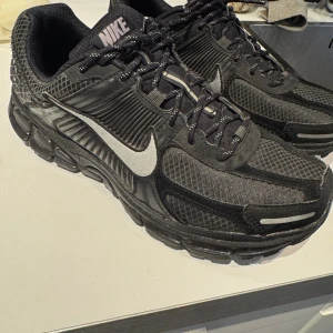 Nike Vomero 5 svart/militärgröna sneakers - Nike Vomero 5 sneakers i svart med vit swoosh. Skorna har mesh och syntetmaterial i ovandelen, snörning och en chunky platt sula. Detaljer i grått och reflekterande inslag på sidan och tungan. Perfekt för dig som gillar sportig och modern stil. Nypris 1849kr