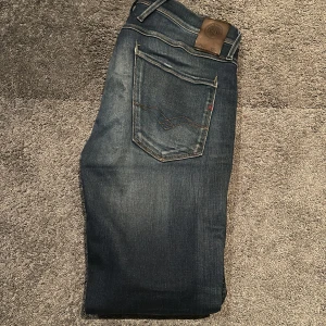 Replay Anbass jeans mörkblå herr - Säljer ett par Replay Anbass jeans i mörkblå tvätt med snygga slitningar och klassiska fem fickor. Jeansen har smal passform, orangea sömmar och läderpatch bak i midjan. Perfekta för dig som gillar en modern och stilren look.