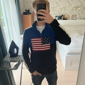 Blå stickad tröja Polo Ralph Lauren - Stickad tröja från Polo Ralph Lauren i blått och svart med vita detaljer på ärmarna. Halv dragkedja och hög krage. Stor amerikansk flagga framtill. Perfekt för dig som vill sticka ut med en klassisk och cool look. Storleken är XL för barn men motsvarar S