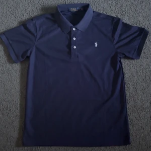 Marinblå pikétröja från Ralph Lauren - Klassisk marinblå pikétröja från Polo Ralph Lauren med vit broderad logga på bröstet. Tröjan har korta ärmar, krage och tre knappar framtill. Perfekt för en stilren och sportig look.