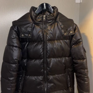 Svart Moncler pufferjacka med huva - Snygg svart pufferjacka från Moncler med glansig finish, huva och dragkedja framtill. Jackan har Moncler-logga på ärmen, praktiska fickor med dragkedja och tryckknappar vid axlarna. Perfekt för kalla dagar när du vill hålla stilen.