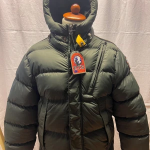 Grön pufferjacka från Parajumpers - Snygg mörkgrön pufferjacka från Parajumpers med huva, dragkedja och coola detaljer som logopatch på ärmen och bröstet. Jackan har en quiltad look och flera praktiska fickor. Perfekt för kalla dagar när du vill hålla stilen.