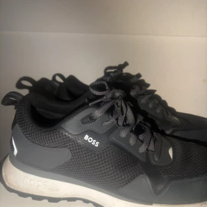 sneakers från BOSS - Snygga svarta sneakers från BOSS med grå detaljer och vit sula. Skorna har meshpaneler för extra ventilation och snörning framtill. Perfekt för dig som gillar stilrena och sportiga skor med modern design. Storlek 44