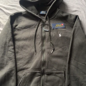 Mörkgrön hoodie från Polo Ralph Lauren - Snygg svart hoodie från Polo Ralph Lauren med dragkedja och huva. Tröjan har snörning vid halsen, klassisk Polo-logga broderad på bröstet och praktiska fickor framtill. Perfekt för chill dagar och streetwear-stil.