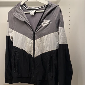 Nike vindjacka grå/svart/vit - Nike vindjacka i storlek 158-170cm med huva och dragkedja. Jackan har färgblock i grått, vitt och svart, samt Nike-logga på bröstet och ryggen. Tillverkad i lätt polyester med meshfoder och praktiska fickor med dragkedja. Perfekt för en sportig streetstil.