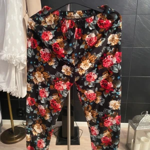 Blommiga svarta byxor med resår - Snygga svarta byxor med färgglatt blommönster i rött, beige och grönt. Byxorna har elastisk midja och rak passform, perfekta för en chill och trendig look. Mjukt och lätt material som känns skönt mot huden.