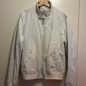 Vit vindjacka från J.Lindeberg XL - Snygg vit vindjacka från J.Lindeberg i storlek X,L men sitter mer som M/L. Jackan har ribbade muddar, ståkrage med knapp, dragkedja framtill och två fickor med knappstängning. Liten broderad logga på bröstet och rutigt foder på insidan. Perfekt för dig som gillar stilren och lite mer vintage design. 