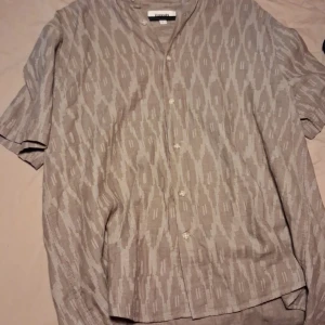 Beige mönstrad henley från Weekday - Säljer en beige henley-skjorta från Weekday med ett unikt geometriskt mönster. Skjortan har korta ärmar, knappar framtill och är tillverkad i ett lätt och luftigt linnematerial. Perfekt för dig som gillar avslappnad och stilren vibe.