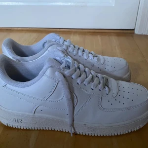 Nike Air Force 1  - Nike Air Force 1 sneakers i helvitt med klassisk låg siluett. Skorna har snörning, perforerad tå och platt sula med tydlig Air-dämpning. Tillverkade i skinn med syntetdetaljer och har rund tå. Perfekt för dig som gillar cleana och tidlösa sneakers.