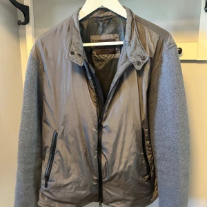 Grå cardigan från Massimo Dutti - Snygg grå cardigan från Massimo Dutti med glansig framsida i syntet och stickade ärmar i en ljusare grå ton. Jackan har ståkrage med tryckknappar, två fickor med dragkedja och hel dragkedja framtill. Perfekt för dig som gillar stilren och modern look.