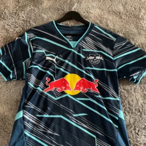 RB Leipzig Openda matchtröja Puma - Snygg RB Leipzig fotbollströja med Openda och nummer 11 på ryggen. Tröjan är marinblå med ljusblå och vita grafiska ränder, Red Bull-logga på bröstet och Puma-logga på axeln. Kortärmad med V-ringad krage, tillverkad i lätt polyester.    Detta är en Replika!