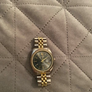 Avery herrklocka i guld och silver - Stilren herrklocka från Avery med rund urtavla i mörkgrått och detaljer i guld. Armbandet är i klassisk länkdesign med både guld- och silverfärgade partier. Klockan har datumvisning och quartz-urverk. Perfekt för dig som gillar tidlös design och metalliska färger.