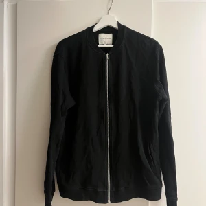 Svart bomberjacka från Studio Total - Svart bomberjacka från Studio Total i storlek L. Jackan har en stilren design med rund hals, hel dragkedja framtill och ribbade muddar vid ärmslut och nederkant. Tillverkad i mjuk bomull, perfekt för en clean och enkel look.