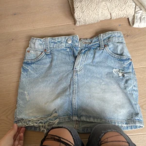 Ljusblå jeanskjol från H&M - Säljer en ljusblå jeanskjol från H&M med slitna detaljer och fransad nederkant. Kjolen har klassisk femficksdesign, bälteshällor och knappstängning framtill. Perfekt för dig som gillar en avslappnad och trendig denimlook.